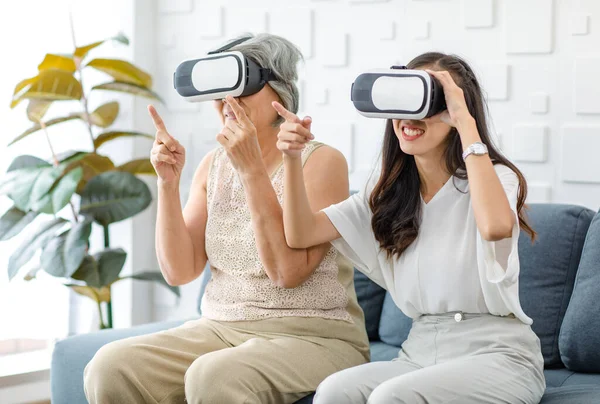 Asian young cheerful daughter wearing VR virtual reality goggles headset sitting smiling on cozy sofa in living room teaching old senior grey hair pensioner mother playing streaming 3d game online.