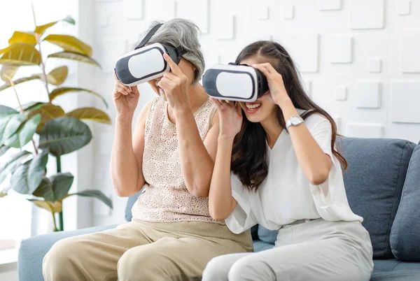 Asian young cheerful daughter wearing VR virtual reality goggles headset sitting smiling on cozy sofa in living room teaching old senior grey hair pensioner mother playing streaming 3d game online.