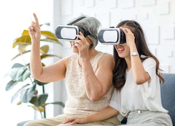 Asian young cheerful daughter wearing VR virtual reality goggles headset sitting smiling on cozy sofa in living room teaching old senior grey hair pensioner mother playing streaming 3d game online.