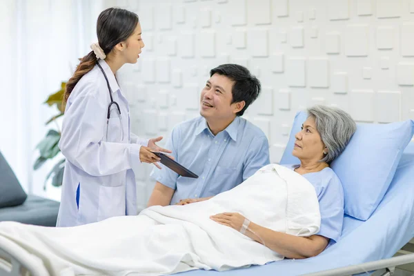 Asian cheerful son holding arm of old senior pensioner mother patient in blue hospital uniform laying down on bed up while professional doctor practitioner in white lab coat visiting in clinical ward.