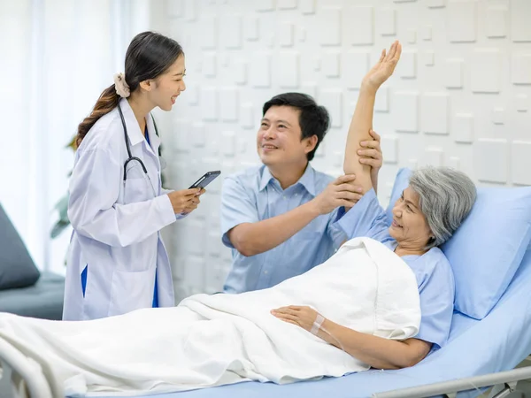 Asian cheerful son holding arm of old senior pensioner mother patient in blue hospital uniform laying down on bed up while professional doctor practitioner in white lab coat visiting in clinical ward.