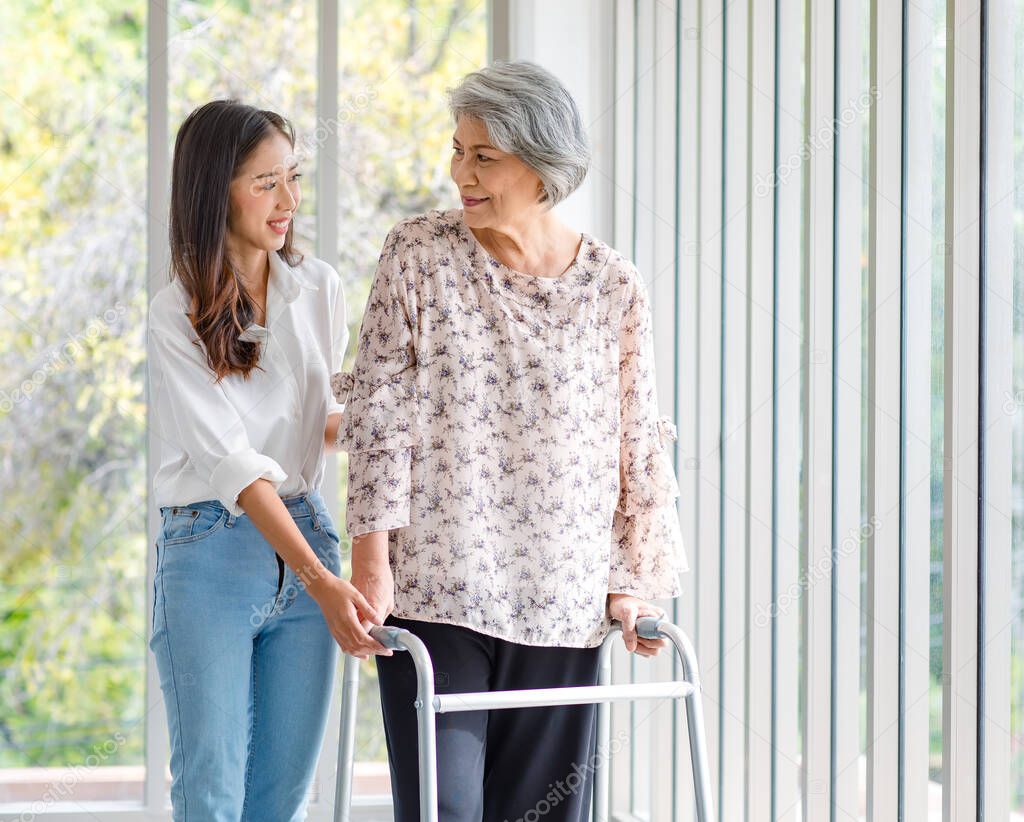 Asian young beautiful happy female granddaughter smiling helping cheerful old senior healthy gray hair retired pensioner grandmother walking using support assist four legged walker in living room.