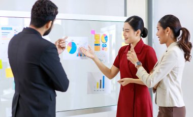 Asyalı genç, güzel, profesyonel, başarılı iş kadını gülümseyip grafik çizelgesini sunuyor. Veri belgelerini cam tahtada kadın ve erkek meslektaşlarına sunuyor..