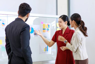 Asyalı genç, güzel, profesyonel, başarılı iş kadını gülümseyip grafik çizelgesini sunuyor. Veri belgelerini cam tahtada kadın ve erkek meslektaşlarına sunuyor..