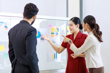 Asyalı genç, güzel, profesyonel, başarılı iş kadını gülümseyip grafik çizelgesini sunuyor. Veri belgelerini cam tahtada kadın ve erkek meslektaşlarına sunuyor..