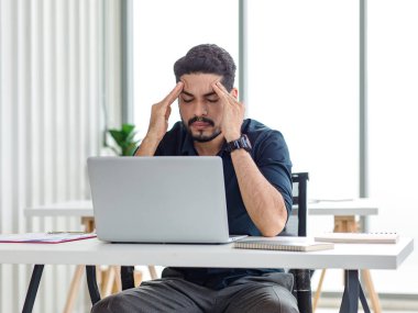 Indian stressed depressed exhausted bearded male businessman sitting at workstation with laptop computer close eyes hold hand on head feeling tired and sleepy after working late overtime last night.