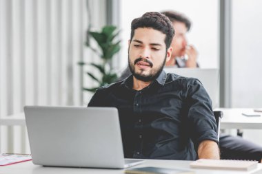 Indian stressed depressed exhausted bearded male businessman sitting at workstation with laptop computer close eyes hold hand on head feeling tired and sleepy after working late overtime last night.