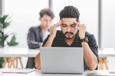 Indian stressed depressed exhausted bearded male businessman sitting at workstation with laptop computer close eyes hold hand on head feeling tired and sleepy after working late overtime last night.