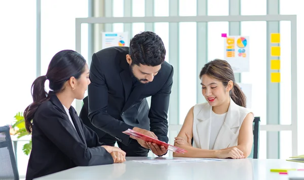 Two Asian young beautiful professional successful businesswoman employees presenting showing paperwork document on clipboard to Indian bearded male businessman manager to approve in meeting room.