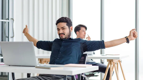 Indian millennial tired exhausted sleepy bearded dull face male businessman sitting at workstation desk with laptop computer stretching arms widely in morning after working late overtime last night.