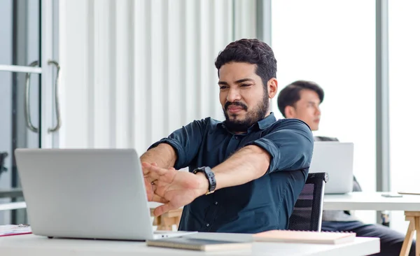 Indian millennial tired exhausted sleepy bearded dull face male businessman sitting at workstation desk with laptop computer stretching arms widely in morning after working late overtime last night.