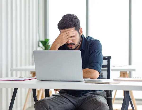 Indian stressed depressed exhausted bearded male businessman sitting at workstation with laptop computer close eyes hold hand on head feeling tired and sleepy after working late overtime last night.