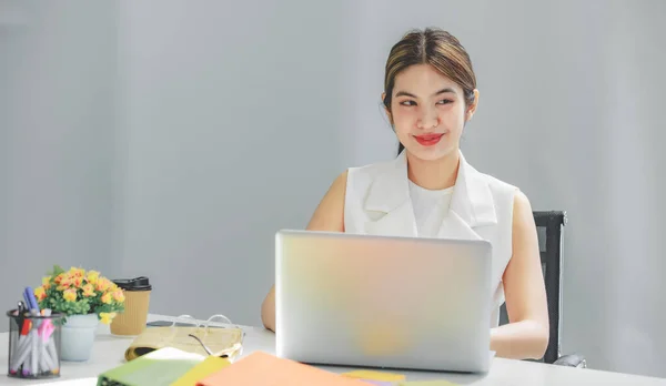 Millennial Asian happy cheerful pretty businesswoman secretary employee sitting smiling enjoy working online typing laptop computer at workstation desk full of document folders in company office.