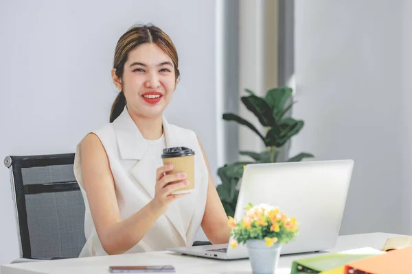 Millennial Asian happy cheerful pretty businesswoman secretary employee sitting smiling enjoy working online typing laptop computer at workstation desk full of document folders in company office.