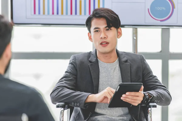 Closeup shot of Asian male businessman intern trainee in casual suit sitting smiling holding tablet in company office.