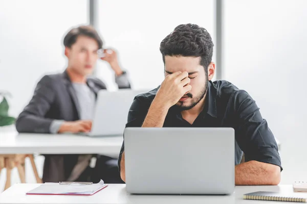 Indian stressed depressed exhausted bearded male businessman sitting at workstation with laptop computer close eyes hold hand on head feeling tired and sleepy after working late overtime last night.