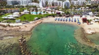 Aerial footage view of Pernera beach in Protaras, Paralimni, Famagusta, Cyprus. The famous tourist attraction golden sandy bay with sunbeds, sun umbrellas water sports, hotels, restaurants, people swimming in sea on summer holidays from above.