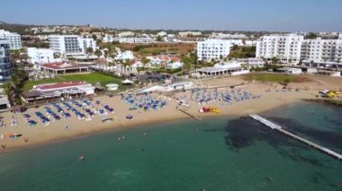 Aerial bird's eye footage view of Sunrise beach at Fig tree in Protaras, Paralimni, Famagusta, Cyprus. The famous tourist attraction family bay with golden sand, sunbeds people swimming sea from above