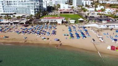 Aerial bird's eye footage view of Sunrise beach at Fig tree in Protaras, Paralimni, Famagusta, Cyprus. The famous tourist attraction family bay with golden sand, sunbeds people swimming sea from above. Drone moving away reveal scene
