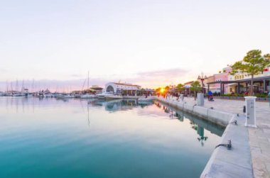 Timelapse footage of sunset at Limassol Marina, Cyprus. People walking fast from dusk to night, city life, yachts docked, a modern landmark for waterfront promenade. Commercial restaurants area view and marina lights.