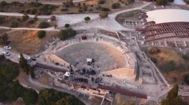 Aerial drone footage of the arcaeological site of the ancient city of Kourio in the district of Limassol, Cyprus. A 360 orbital view of the theatre forming a semicircle,greek roman ruins, beach and fields at sunset from above.
