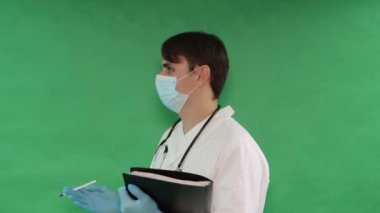 Elinde dosya olan beyaz önlüklü bir doktor inançsızlıkta farklı yönlere döner, yeşil arka planda izole bir şekilde etrafına bakar. Anahtar için yeşil ekran krom anahtar arkaplanı.