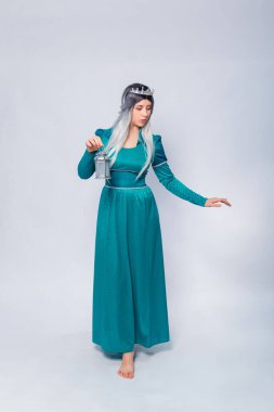 Orta çağ, fantezi, turkuaz bir elbise ve gümüş bir taç giymiş bir prensesin uzun bir portresi. Beyaz bir arka planda, elinde lambayla poz veriyor..