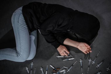 Uyuşturucu bağımlılığı ve aşırı doz konsepti. Kapüşonlu ve kot pantolonlu bir kız elinde şırıngayla karanlık bir odada, dağınık şırıngaların yanında yatıyor..