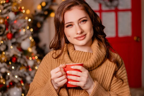 クリスマスツリーを背景に 赤いマグカップを手にした暖かいセーターを着た魅力的な若い女性のクローズアップ肖像 女の子は居心地の良い新年のインテリアでドリンクを飲みます — ストック写真
