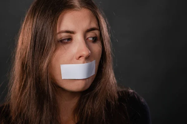 Close-up portrait of a young woman with duct tape sealed in her mouth, restriction of freedom of speech and censored and forbidden to speak and express her opinion, isolated on a dark background.