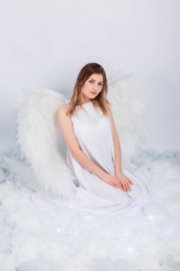 Beyaz ceketli, arkasında büyük beyaz kanatları olan çekici bir kız beyaz bulutlarda otururken poz veriyor..