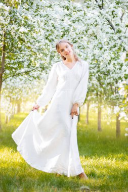 Çiçek açan elma ağaçlarının arka planında beyaz elbiseli genç bir kadın. Kız, bahar parkında çiçeklerin arka planında poz veriyor..