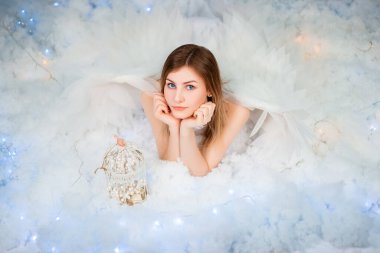 An attractive girl in a white tunic with large white wings behind her back poses while lying in white luminous clouds.