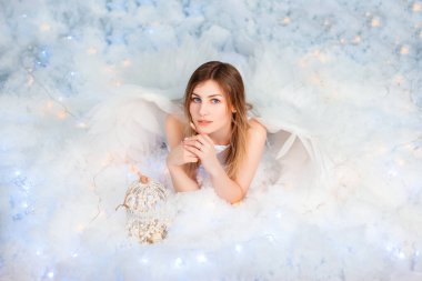 An attractive girl in a white tunic with large white wings behind her back poses while lying in white luminous clouds.