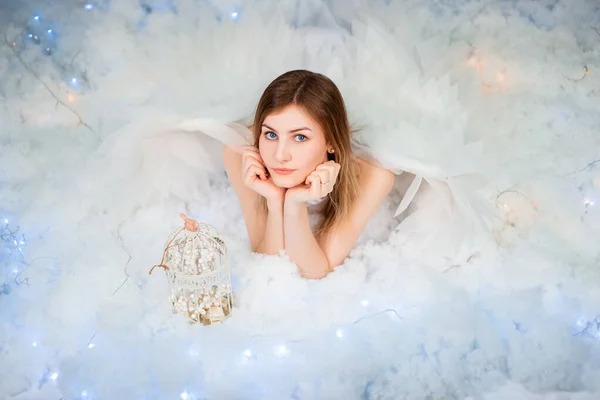 An attractive girl in a white tunic with large white wings behind her back poses while lying in white luminous clouds.