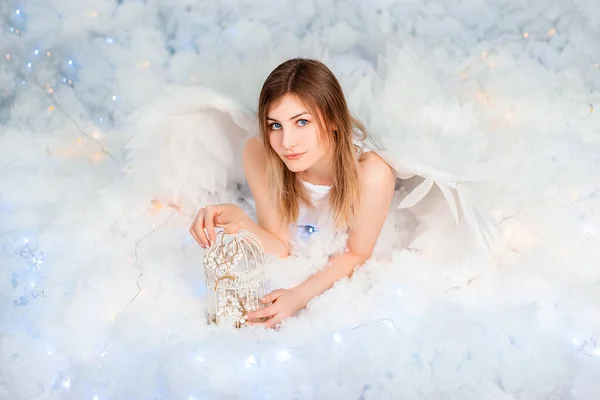 An attractive girl in a white tunic with large white wings behind her back poses while lying in white luminous clouds.