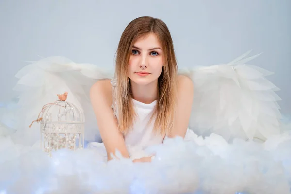 An attractive girl in a white tunic with large white wings behind her back poses while lying in white luminous clouds.