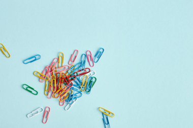 Açık mavi arkaplanda çok renkli ataçlar var. Zımbalar çerçevenin sol alt kısmında rastgele sıralanmış. Yukarıdan görüntüle.