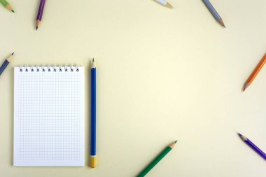 Not defteri ve kalem sarı arka planda yatıyor. Eşyalar sol alt köşede yer alıyor. Renkli kalemler çarşafın kenarlarında belirli bir sırayla durmazlar. Yukarıdan bak, düz yat
