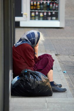 Evsiz bir kadın bir binanın yanında yerde oturuyor. Yandan bakınca, kadının yüzü görünmüyor..