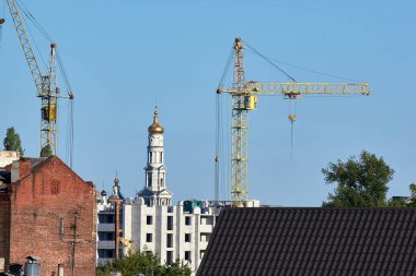 Çok katlı bir binanın inşaatı. Çatı ve eski evin bir kısmı ön planda görünüyor. Arka planda kiliselerin kubbeleri görülüyor..