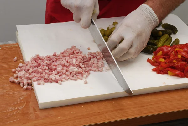 Cortar pequeños trozos de salchicha. Corte de salchichas, pepino y pimienta en pizarra blanca . — Foto de Stock