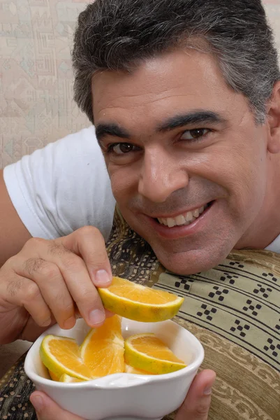Homem adulto médio comendo fatias de laranja em um sofá . — Fotografia de Stock