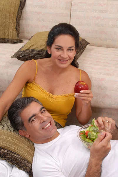 Casal adulto médio comendo salada e frutas em uma sala de estar . — Fotografia de Stock