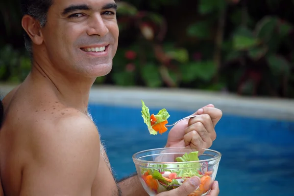 Young man eating vegetables behind a swimming pool. — Stock Photo, Image