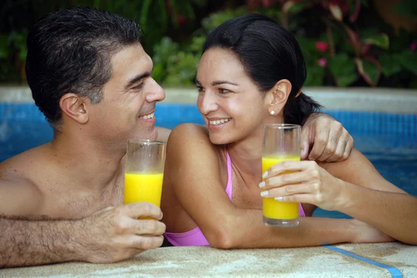 Jovem casal latino bebendo suco de laranja em uma piscina . — Fotografia de Stock