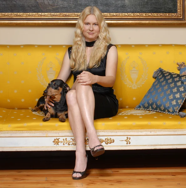 Młoda blond kobieta z mały pies siedzi na elegancki kanapa żółty. — Zdjęcie stockowe