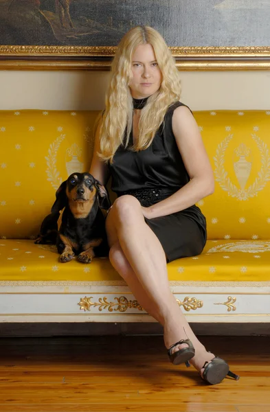 Young blonde woman with a little dog sitting on an elegant yellow sofa. — Stockfoto