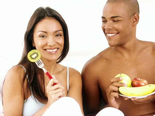 Jonge Spaanse paar genieten van en het eten van fruitsalade op bed. — Stockfoto