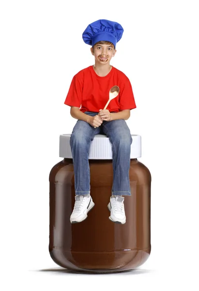 Bambino cuoco seduto sopra una pentola di cioccolato, bambino che tiene un cucchiaio di cioccolato, bambino che mangia crema di cioccolato . — Foto Stock